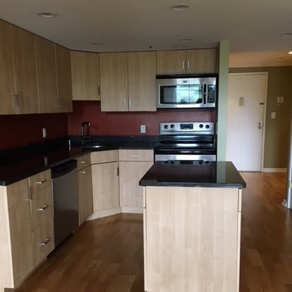 Modern kitchen with light wood cabinets, black countertops, stainless steel appliances, and island