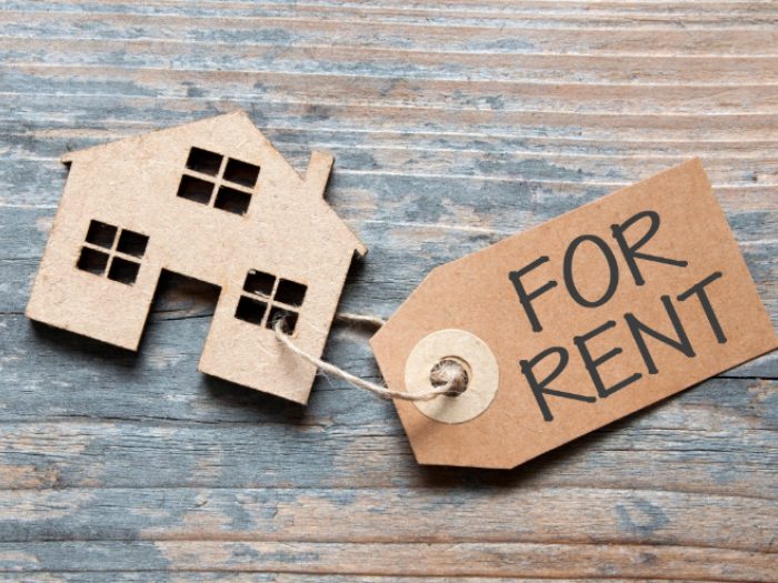 Cardboard house model with "FOR RENT" tag on wooden surface