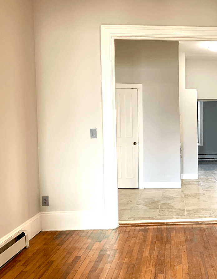 Empty room with hardwood flooring, beige walls, white trim, and doorway to tiled kitchen area