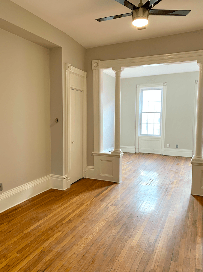 Empty living room with hardwood floors, cream walls, ceiling fan, and columns separating rooms