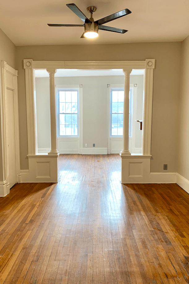 Empty room with hardwood floors, ceiling fan, and white columns framing doorway to sunlit room