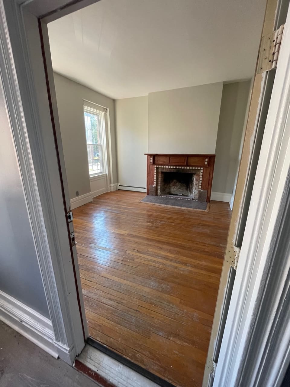 Empty living room with hardwood floors, fireplace with wooden mantel, and window with natural light