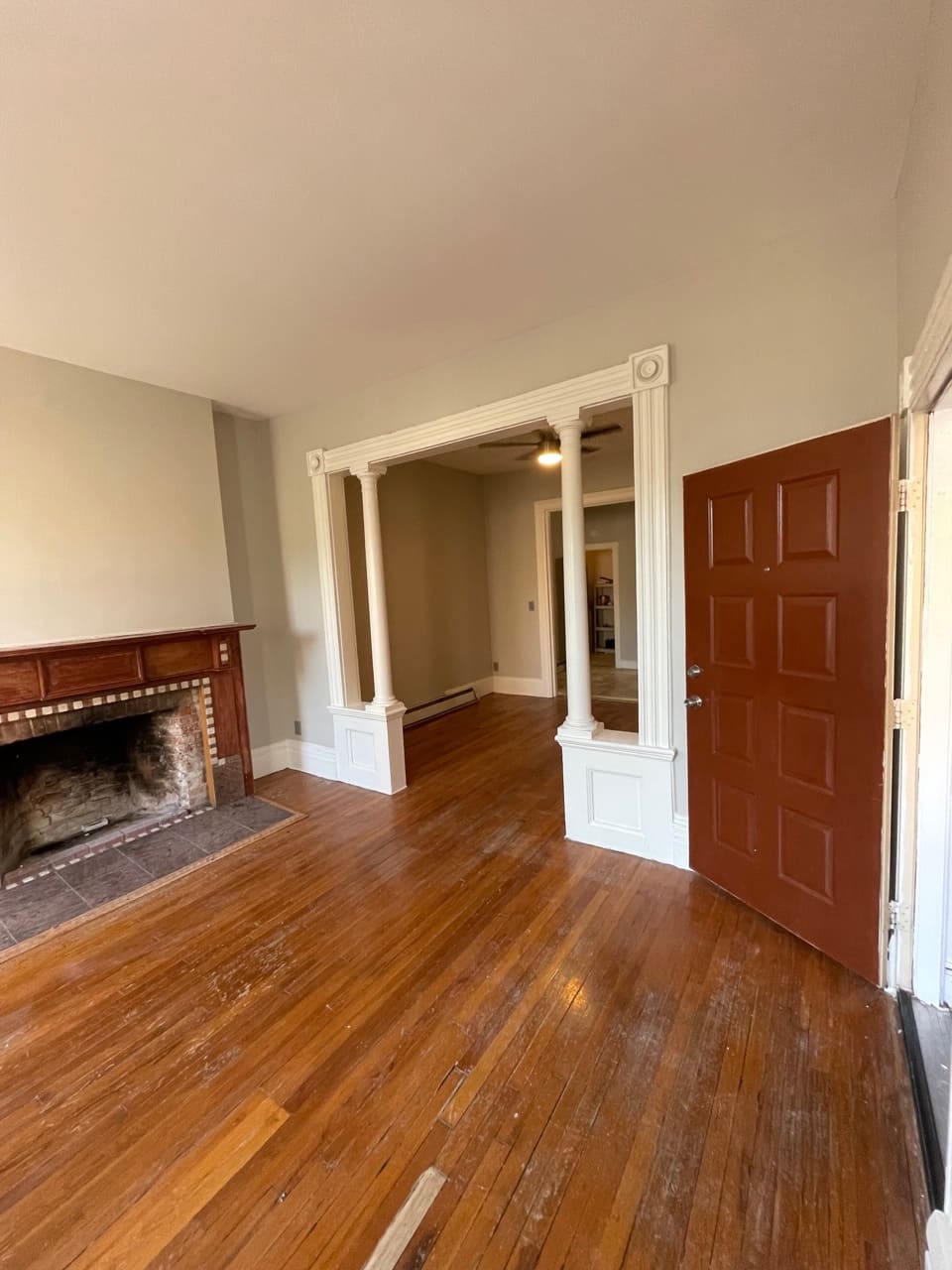 Empty living room with fireplace, hardwood floors, white columns, and red door