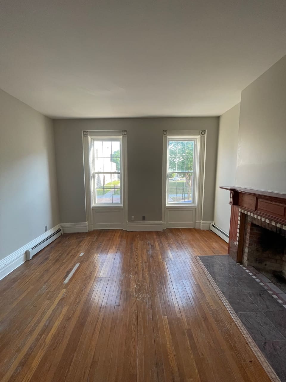 Empty living room with hardwood floors, fireplace, and two tall windows overlooking outdoor area