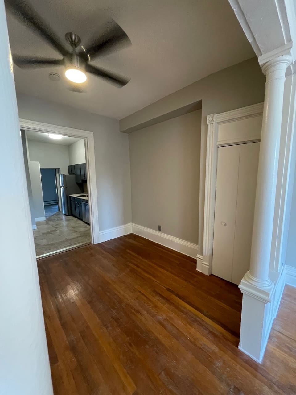 Empty apartment hallway with hardwood floors, ceiling fan, and kitchen visible through doorway