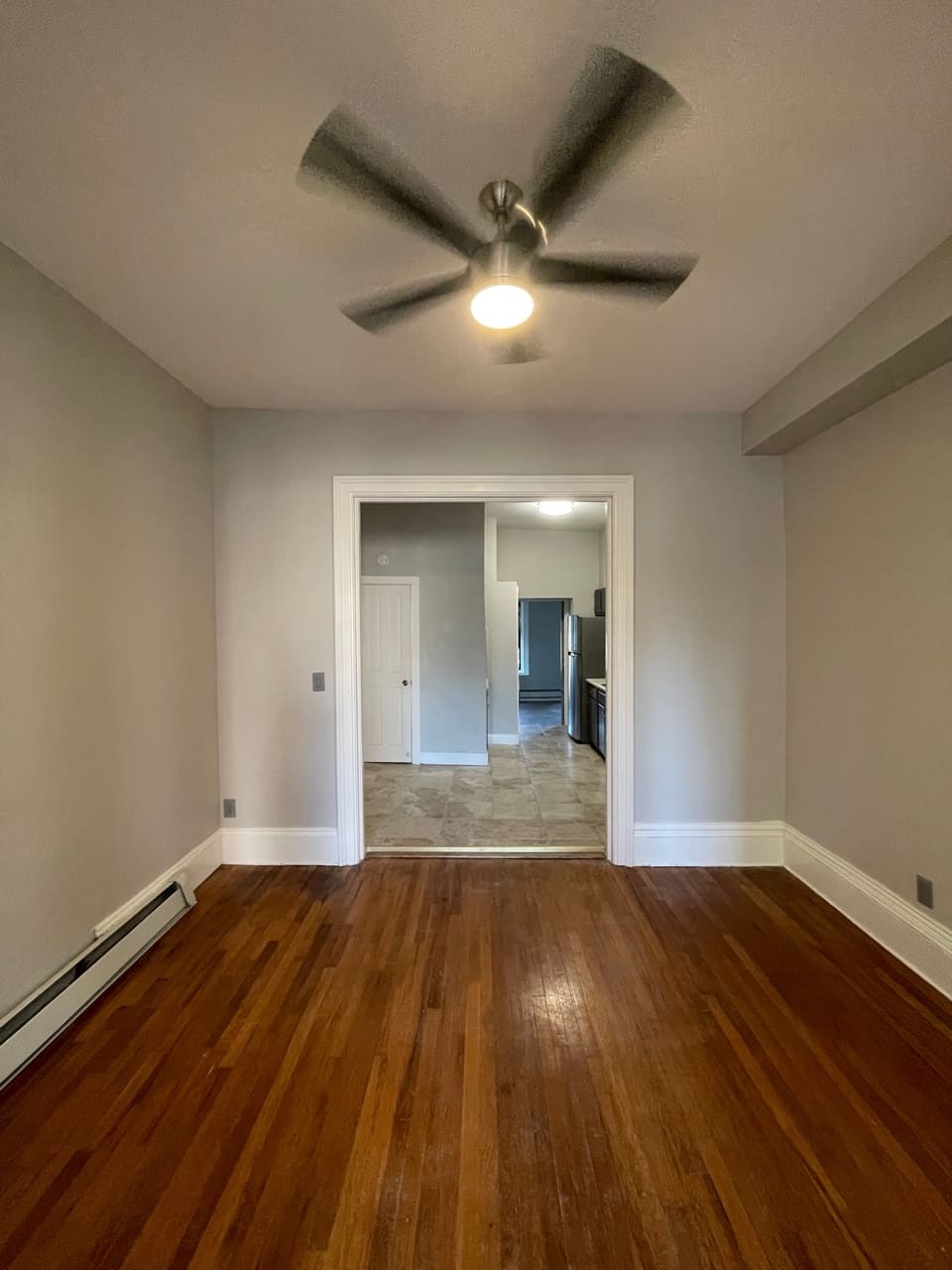 Empty room with hardwood floor, ceiling fan, and open doorway leading to kitchen area